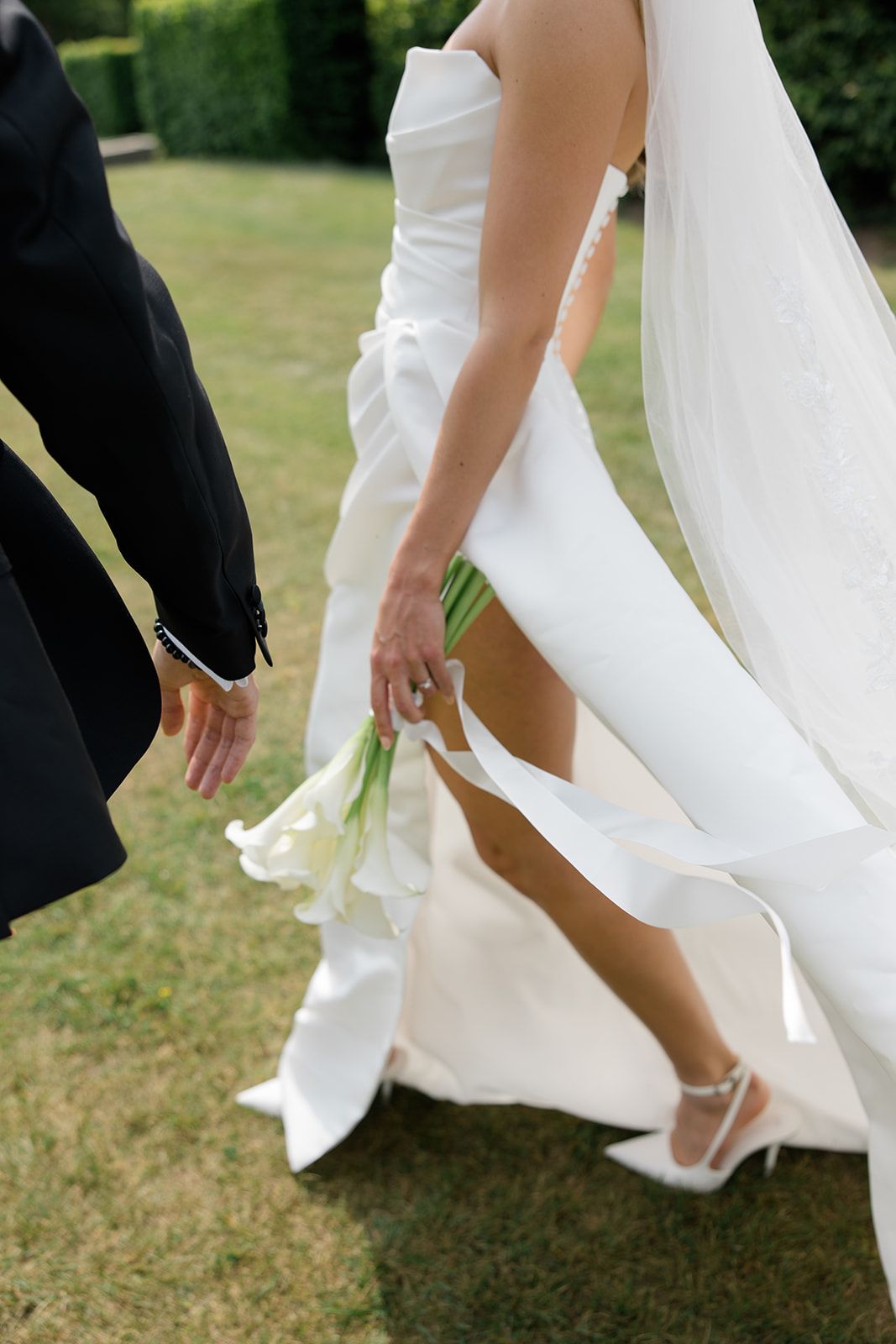Close-up view of bride in white wedding dress holding calla lily bouquet while standing on grass with groom beside her.