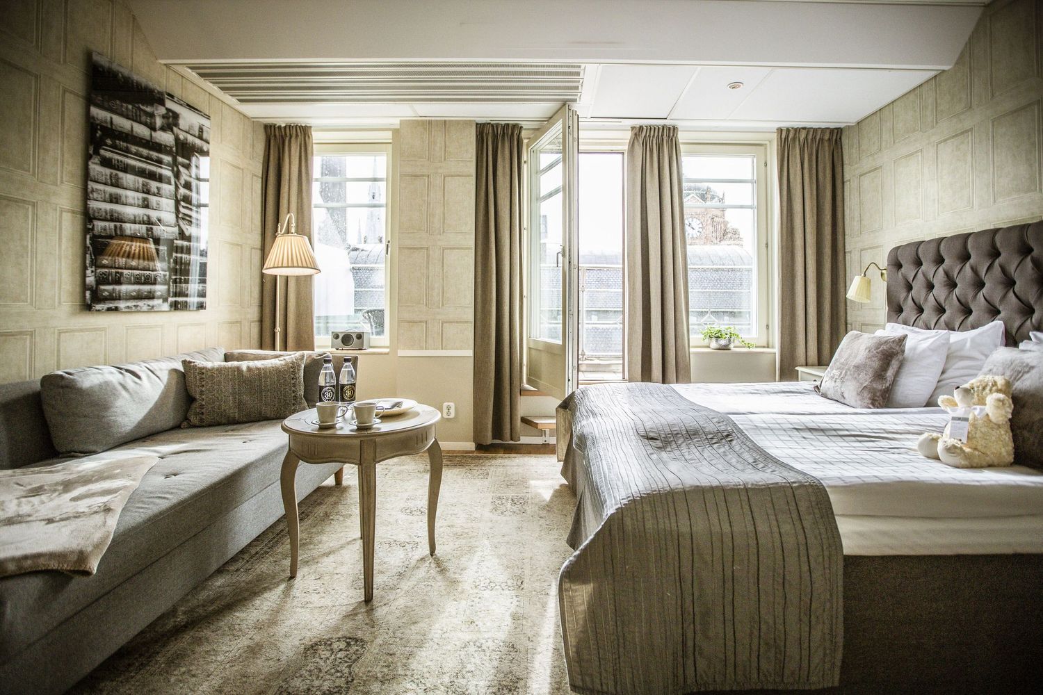 Elegant hotel room interior with tufted headboard, floor-to-ceiling windows, and neutral decor in beige and gray tones.