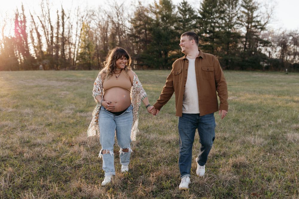 Expectant couple walking together in a field at sunset while holding hands. Virginia/ North Carolina Maternity Session