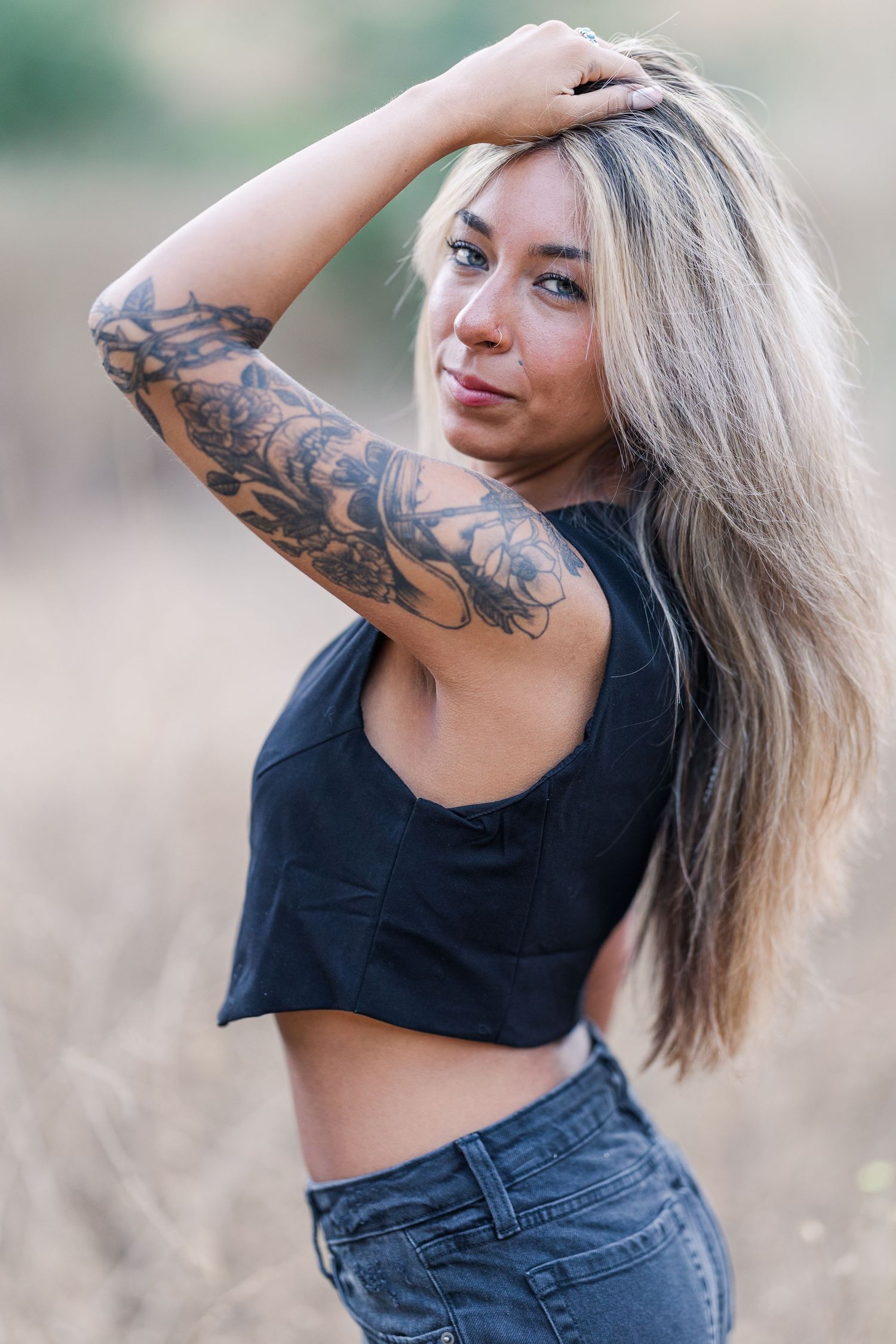 Woman in black crop top and jeans poses outdoors showing ornate floral arm tattoo sleeve in natural lighting.