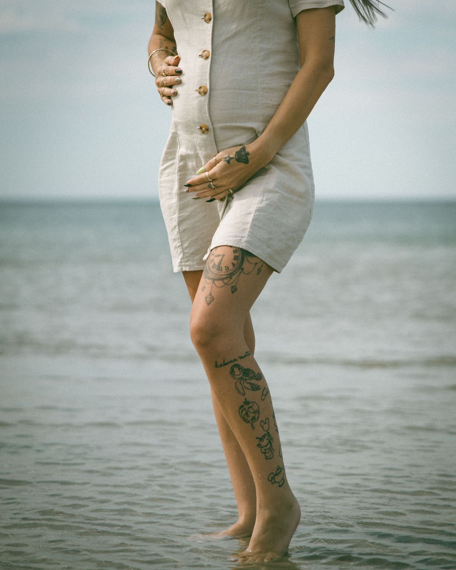 A woman in a white button-up dress stands ankle-deep in ocean water, showing ornate leg tattoos at sunset.