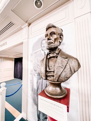 Bronze bust sculpture displayed on red pedestal against white wall in interior hallway setting.