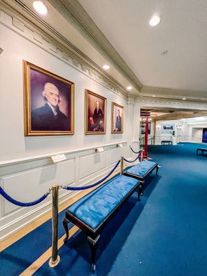 Art gallery corridor with blue carpet, gold-framed portraits on beige walls, and stanchions with velvet ropes.