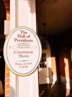 A sign for 'The Hall of Presidents' attraction with continuous shows displayed on a white column against a brick wall.