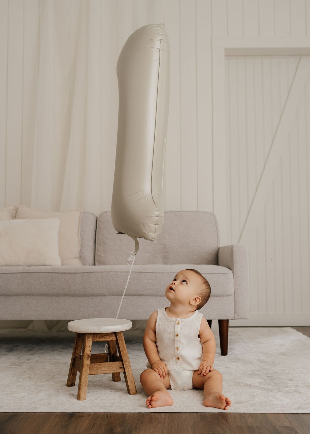 Baby in white romper sitting on floor gazing up at large silver balloon tied to wooden stool.