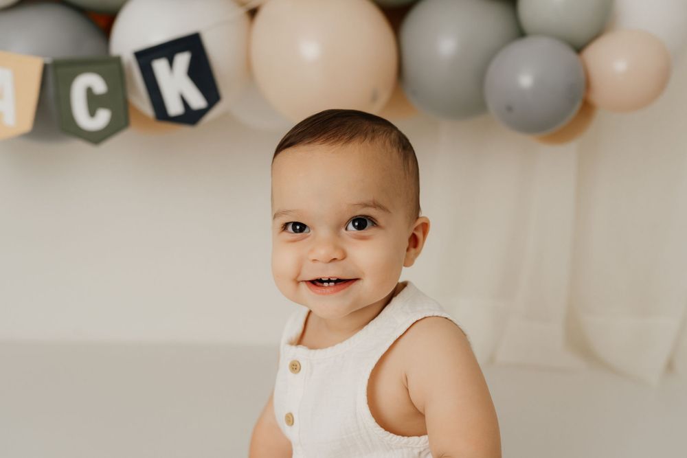 First birthday portrait with decorative balloon garland in neutral colors.