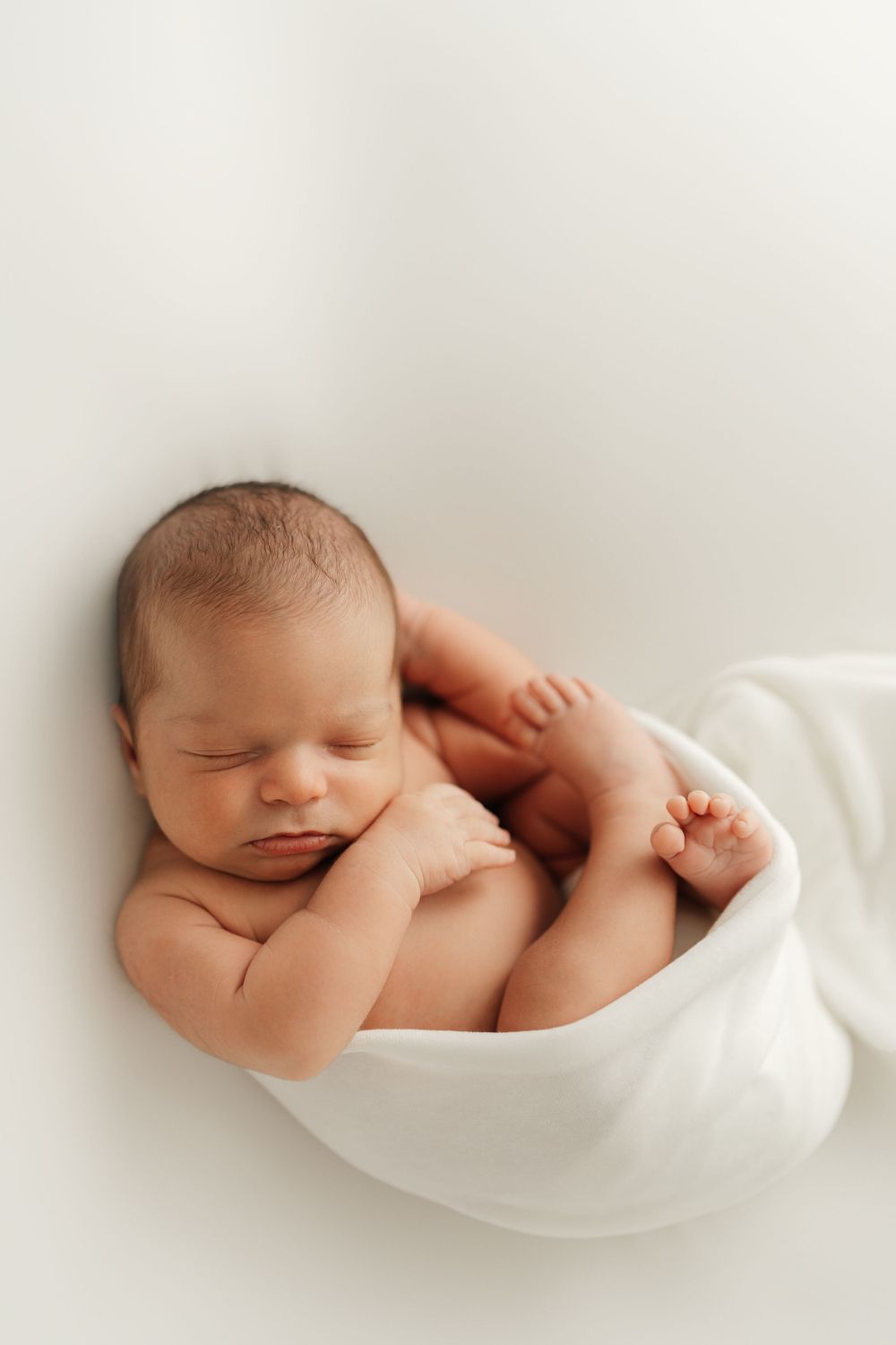 Newborn baby sleeping peacefully while wrapped in soft white fabric against light background.