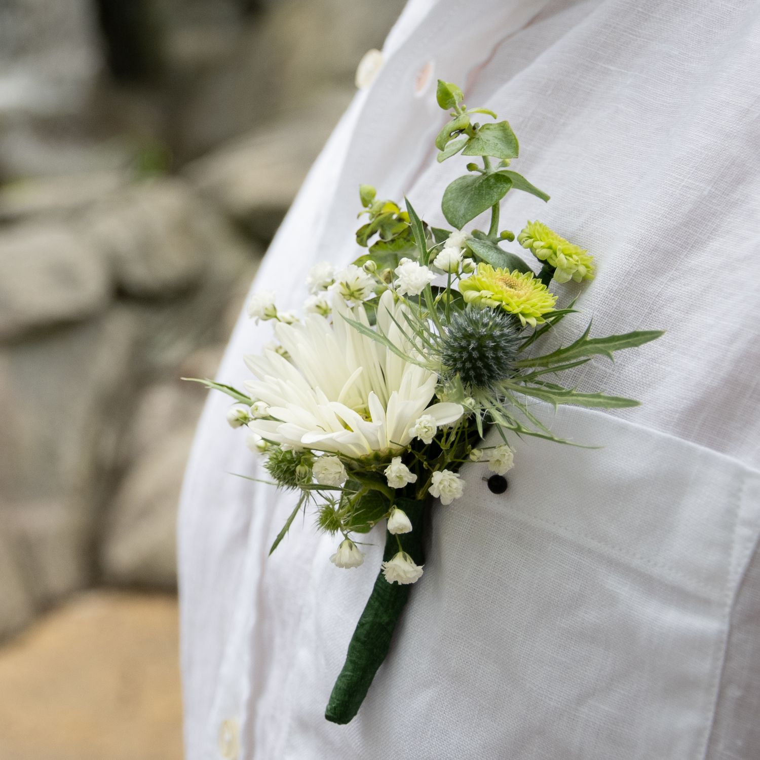 A white chrysanthemum boutonniere with small yellow flowers and delicate greenery pinned to a white dress shirt.