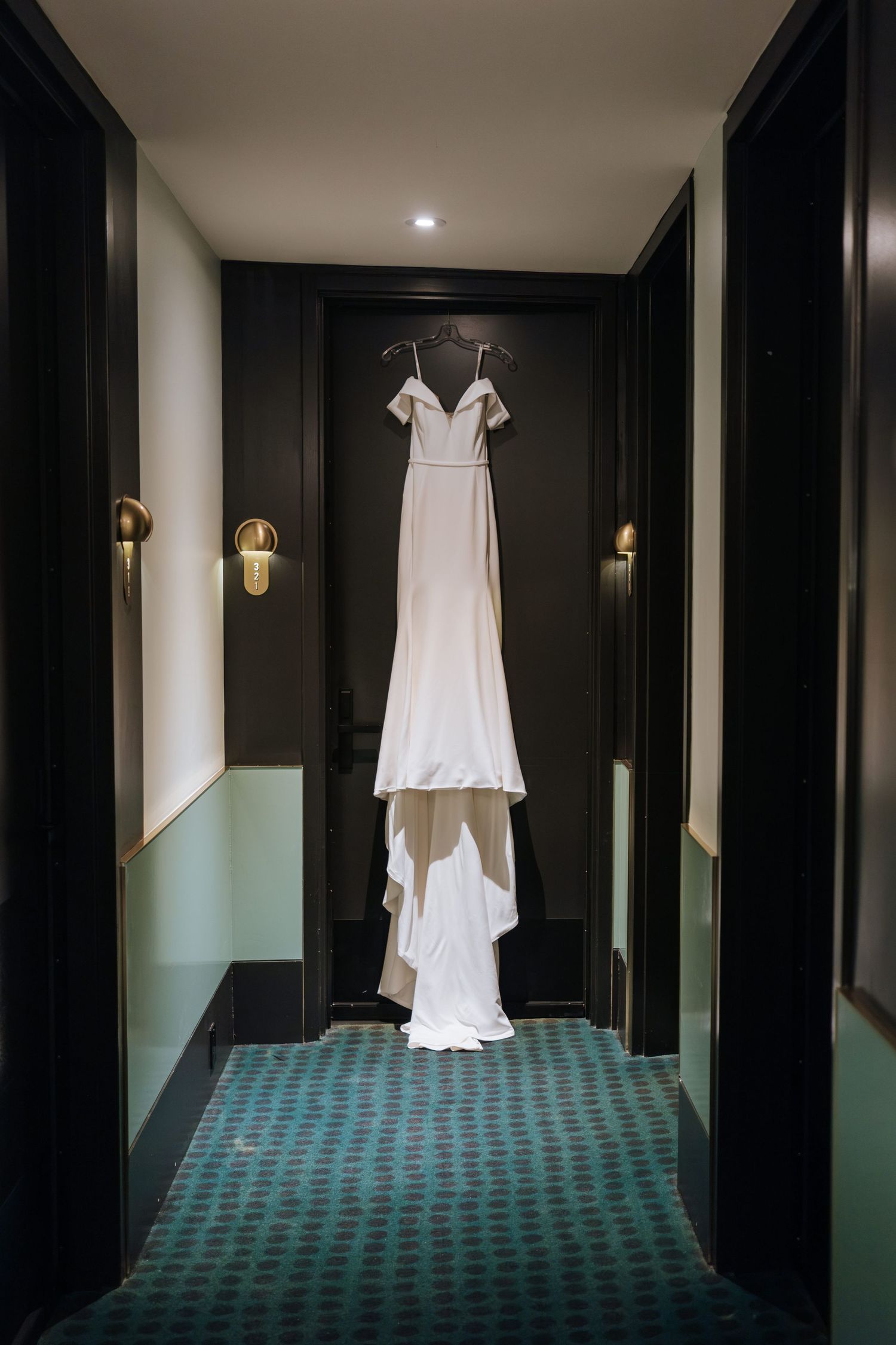 An elegant white wedding dress hanging in a dark hallway with gold sconces.