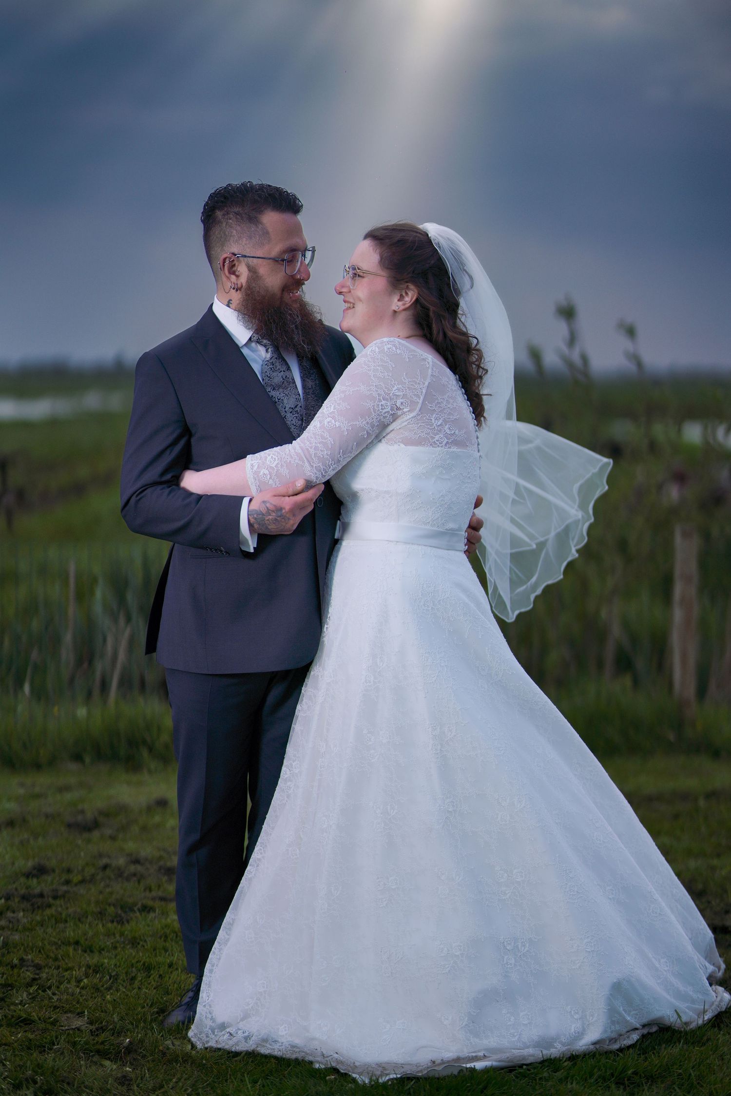 Newlywed couple embracing outdoors in wedding attire, bride in white lace gown with veil, groom in gray suit, dramatic sky.
