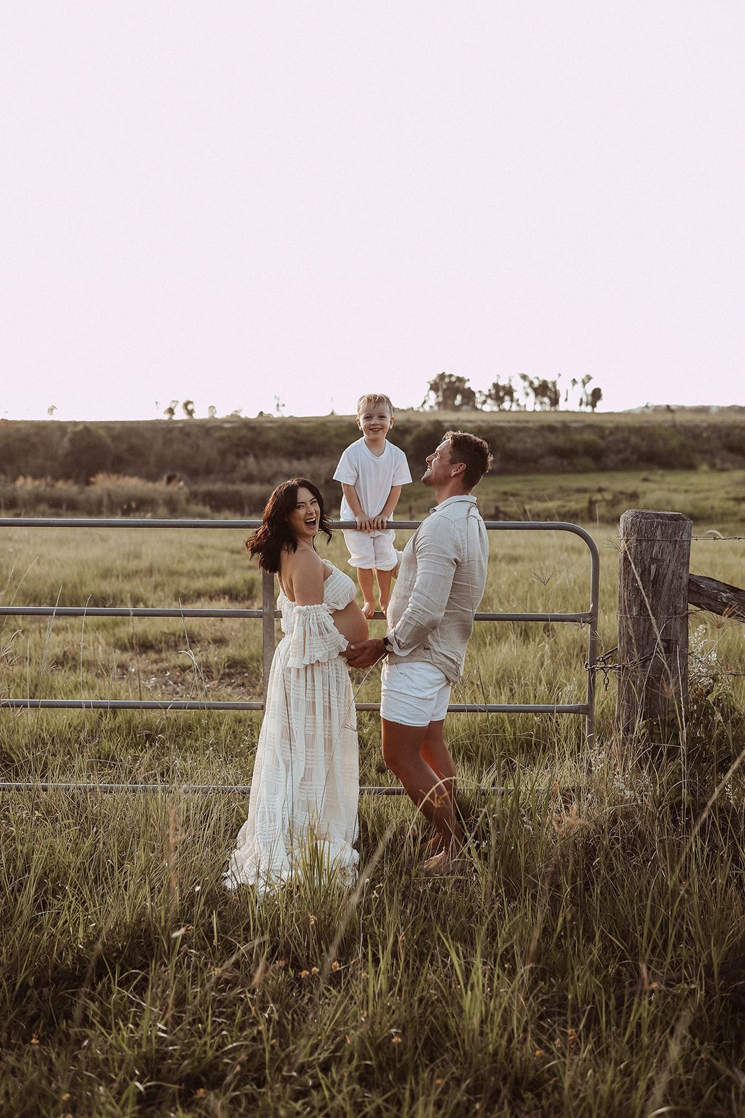 Family of three posing by rustic fence in countryside during sunset.