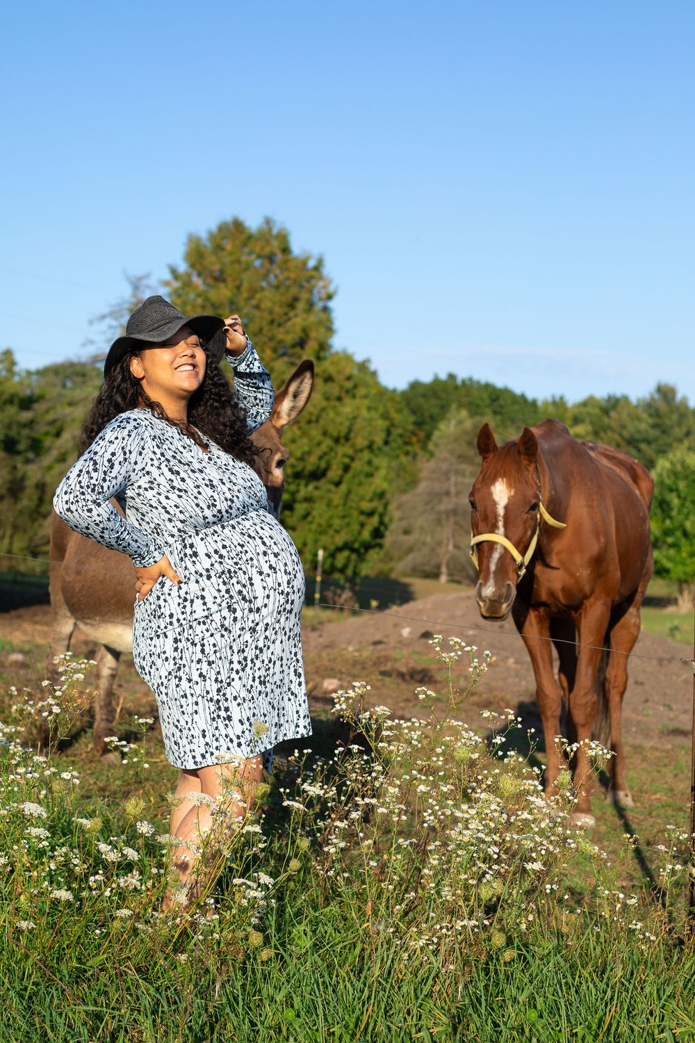Pregnant woman in floral dress and cap poses outdoors with a donkey and brown horse in a sunny field.