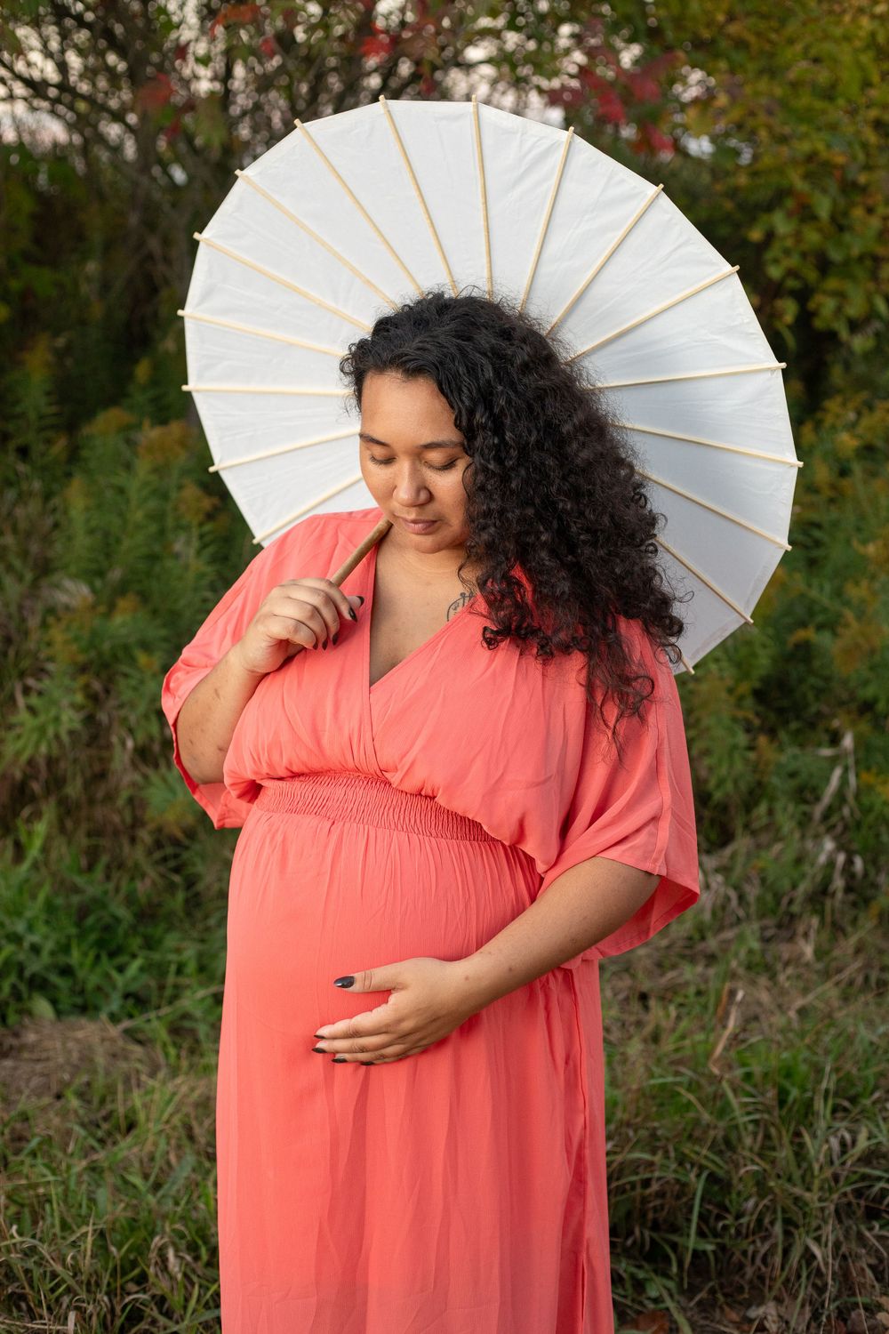 A pregnant woman in a coral dress holds a white parasol while standing in a garden at sunset.