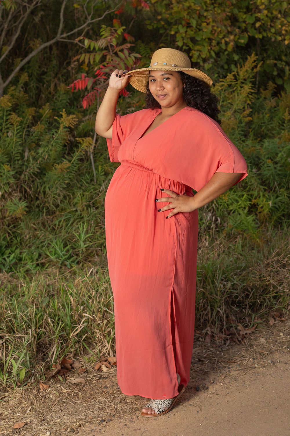 Woman in coral maxi dress and straw hat poses on a nature trail surrounded by autumn foliage.