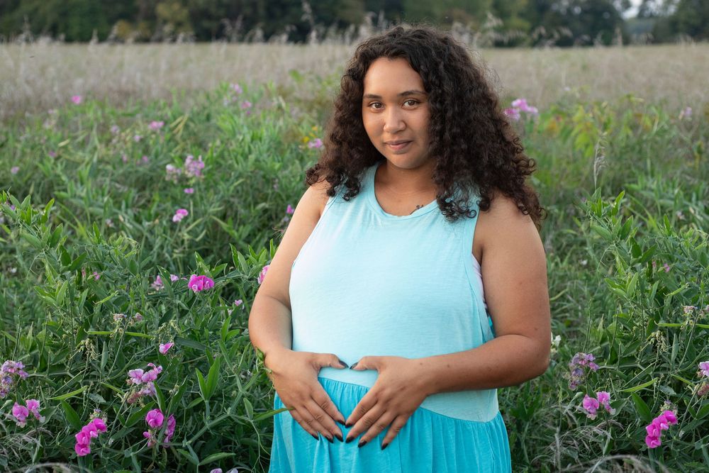 Pregnant woman in teal dress making heart shape over belly, standing in field of pink wildflowers.