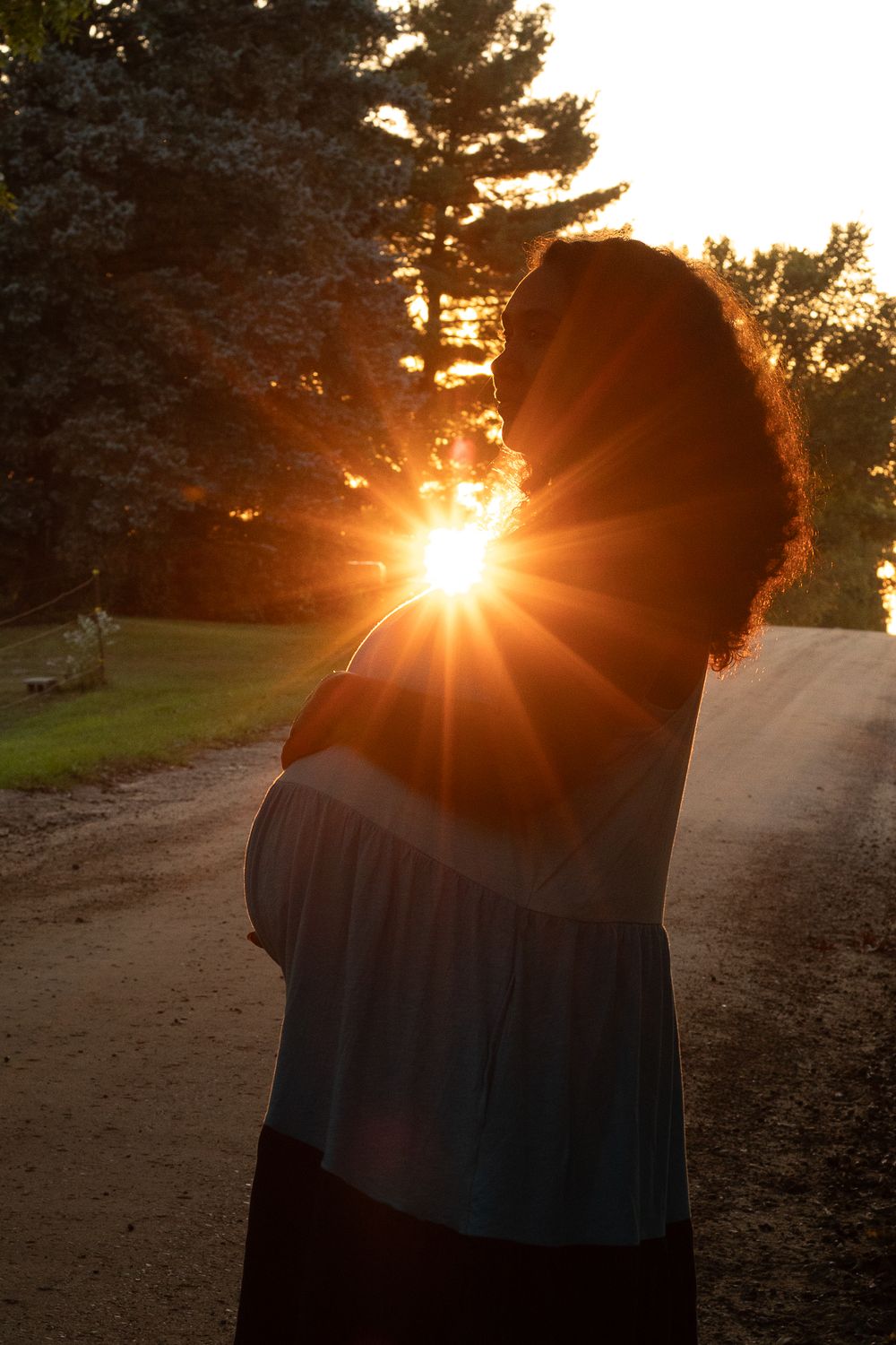 Pregnant woman silhouetted against golden sunset on a rural gravel road, sun rays bursting through, trees in background.