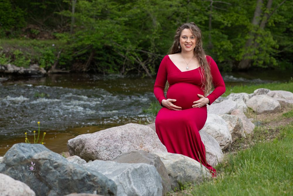 Pregnant woman in red dress sitting on rocks by a river, smiling and cradling her baby bump outdoors.
