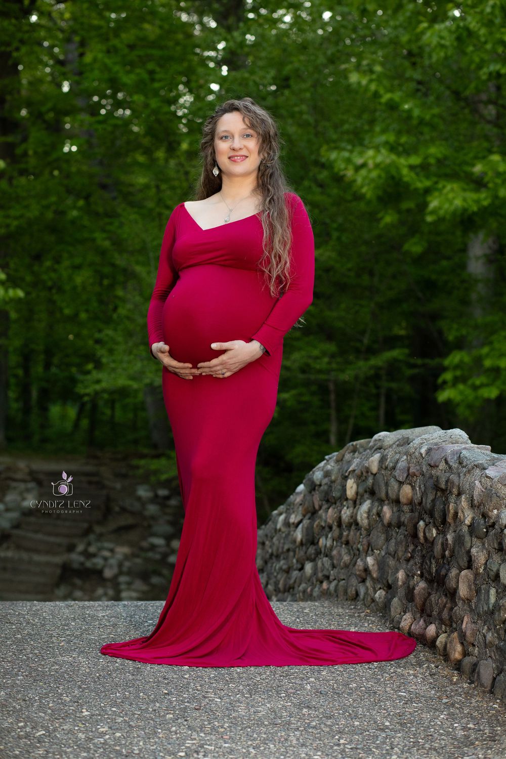 A pregnant woman in a flowing red maternity gown stands beside a stone wall with lush green trees in the background.