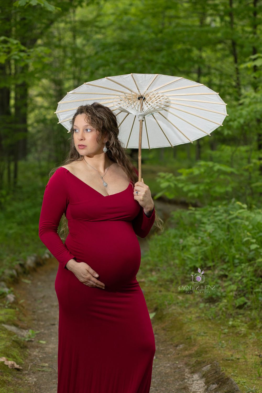 Expectant figure in a flowing red dress holds a white parasol while standing on a forest path surrounded by lush greenery.
