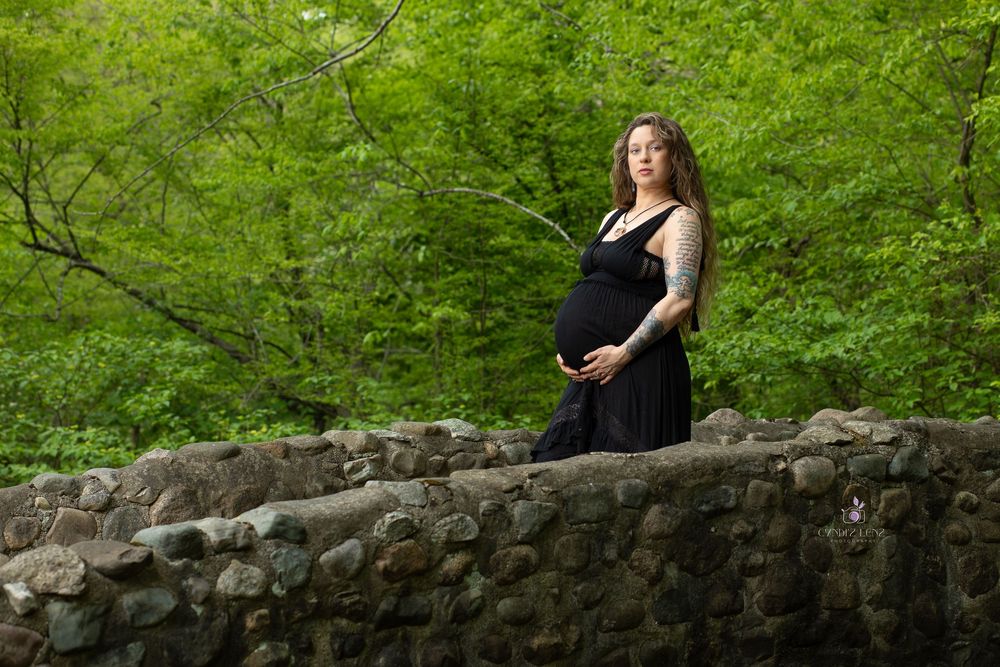 Pregnant woman in black dress standing on stone bridge surrounded by lush green forest trees.