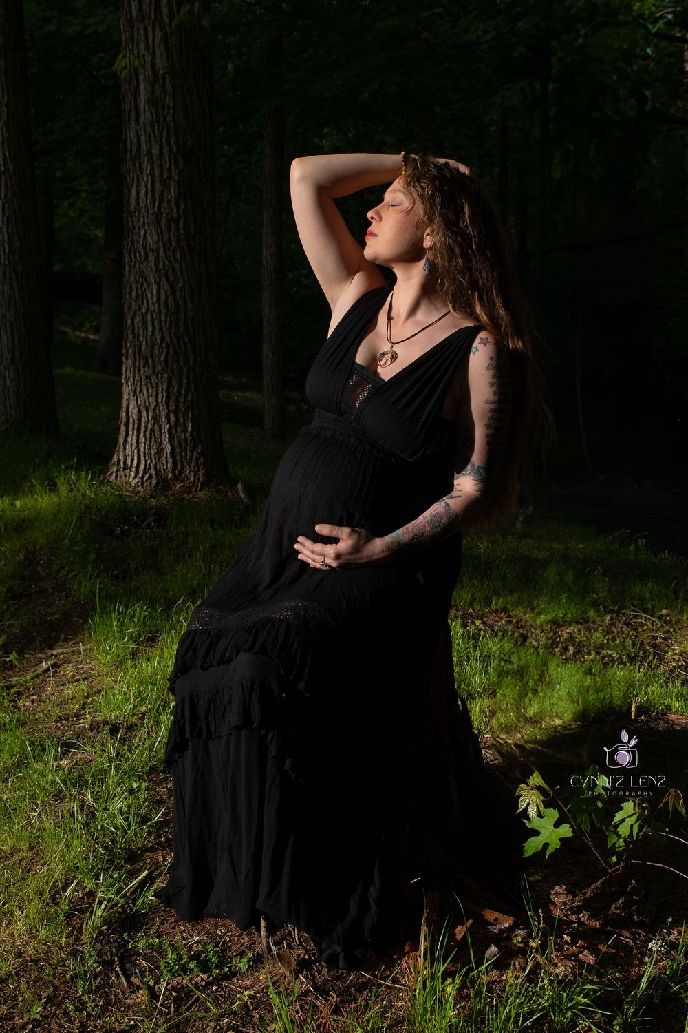 Pregnant woman in black dress poses gracefully in sunlit forest, showcasing tattoos and maternity elegance.