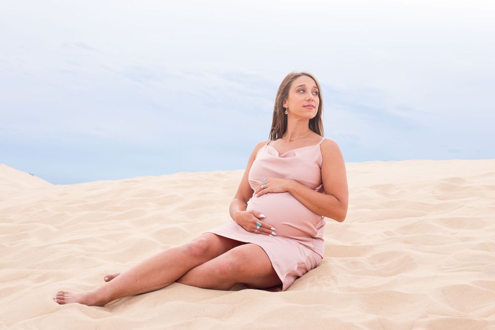 A pregnant woman in a pink dress sits peacefully on sand dunes against a bright blue sky.