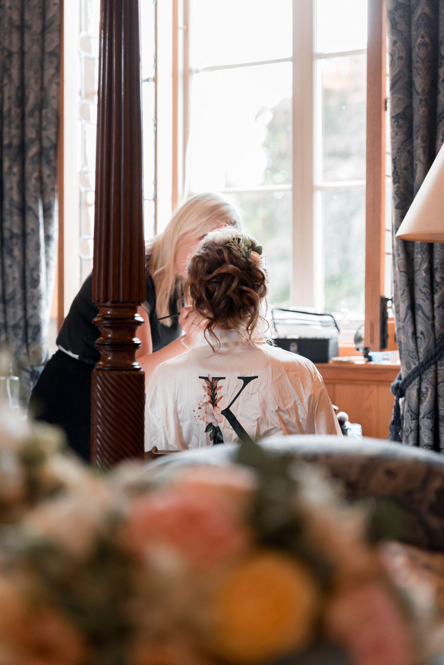 A person in a white monogrammed robe sits at a dressing table near a window with dark curtains and floral arrangements.