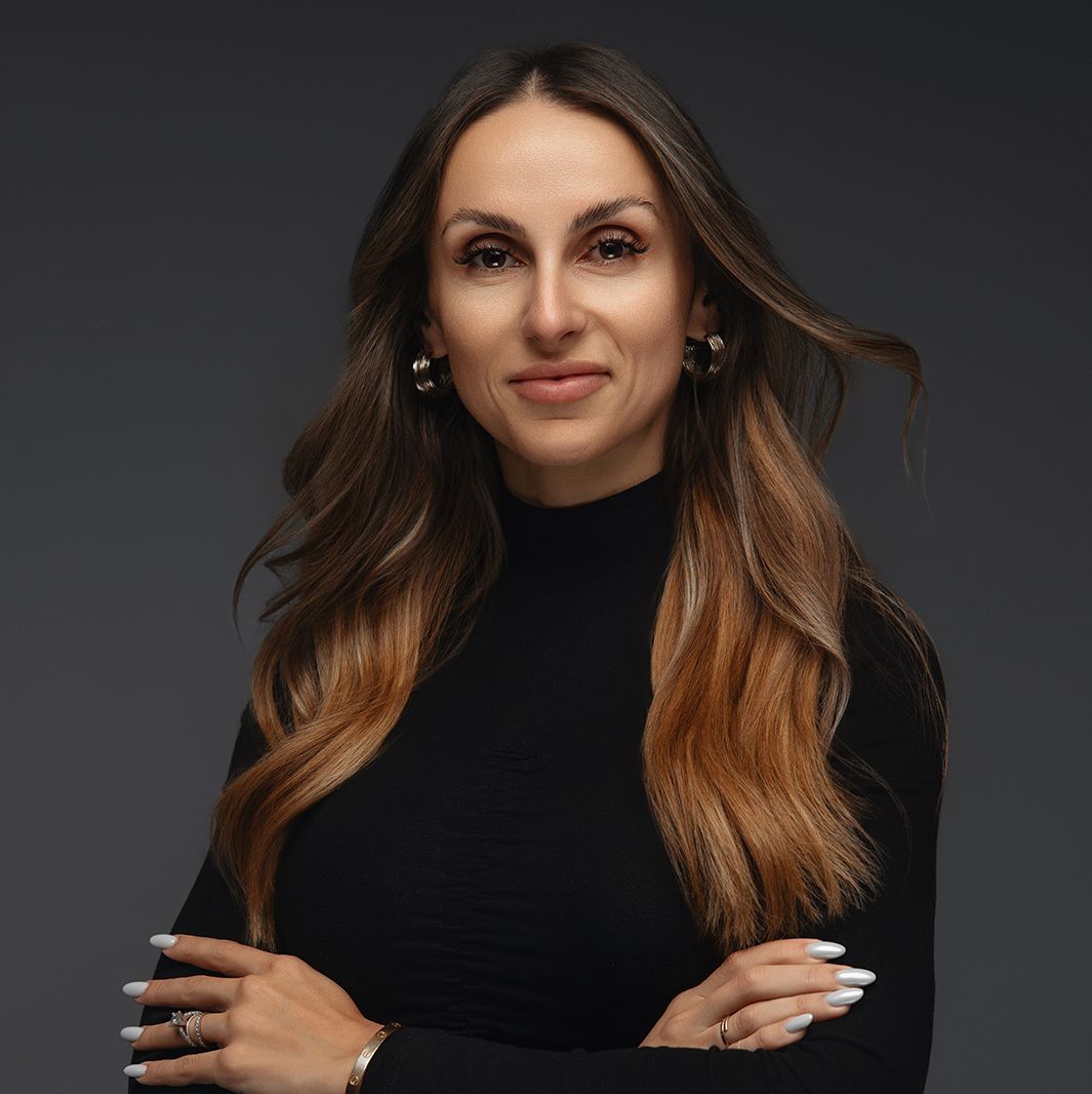 Professional headshot of a person with long wavy brown hair wearing a black turtleneck sweater against dark background.