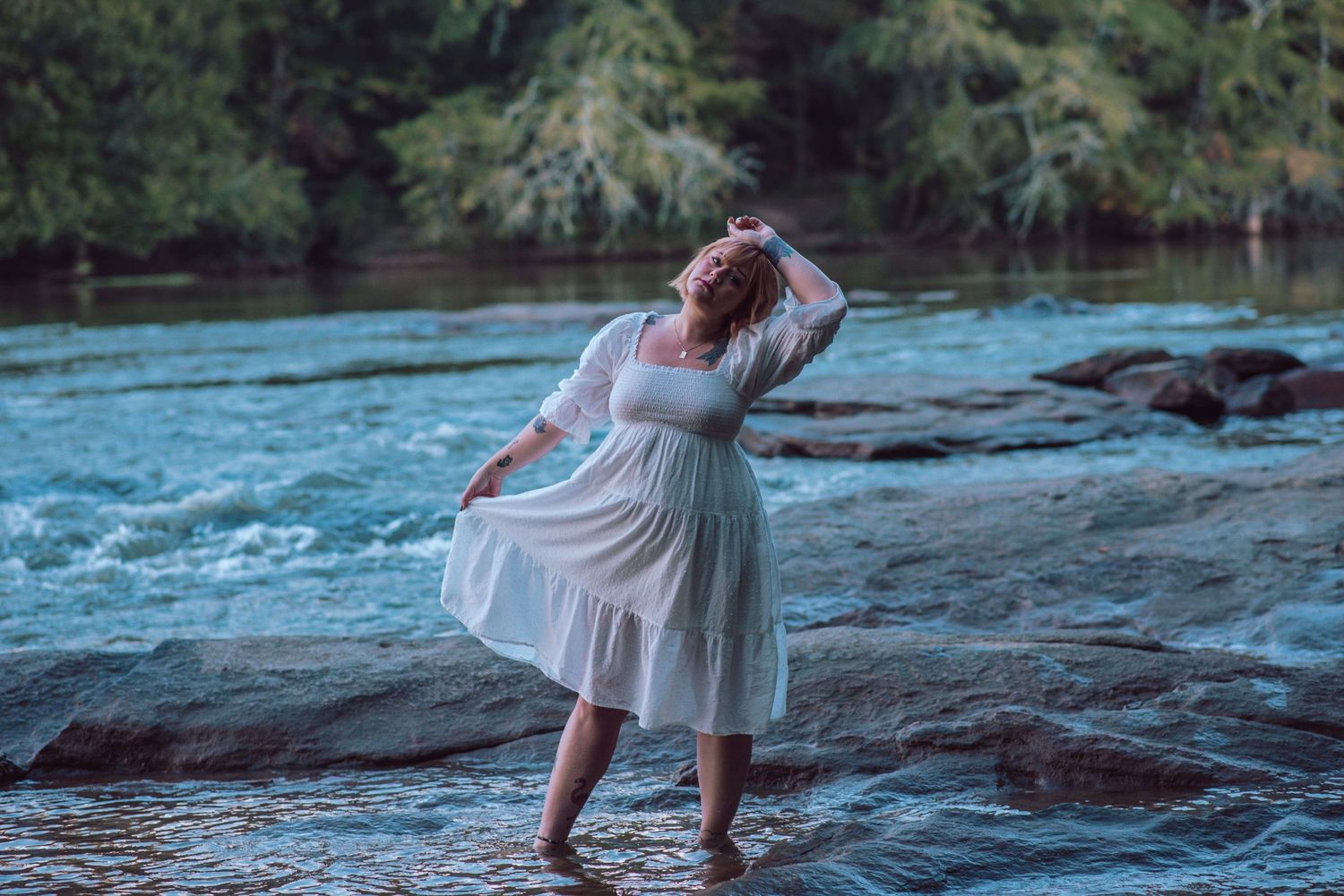 A woman in a flowing white dress stands on rocky river banks during a dreamy photoshoot.
