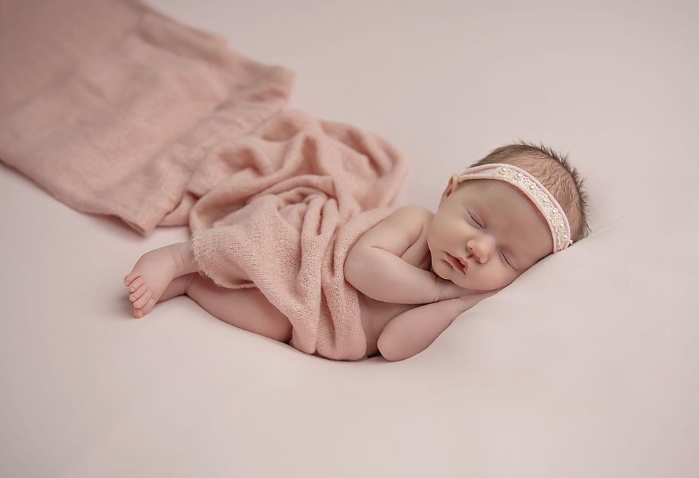 Sleeping newborn wearing headband and wrapped in soft pink blanket.
