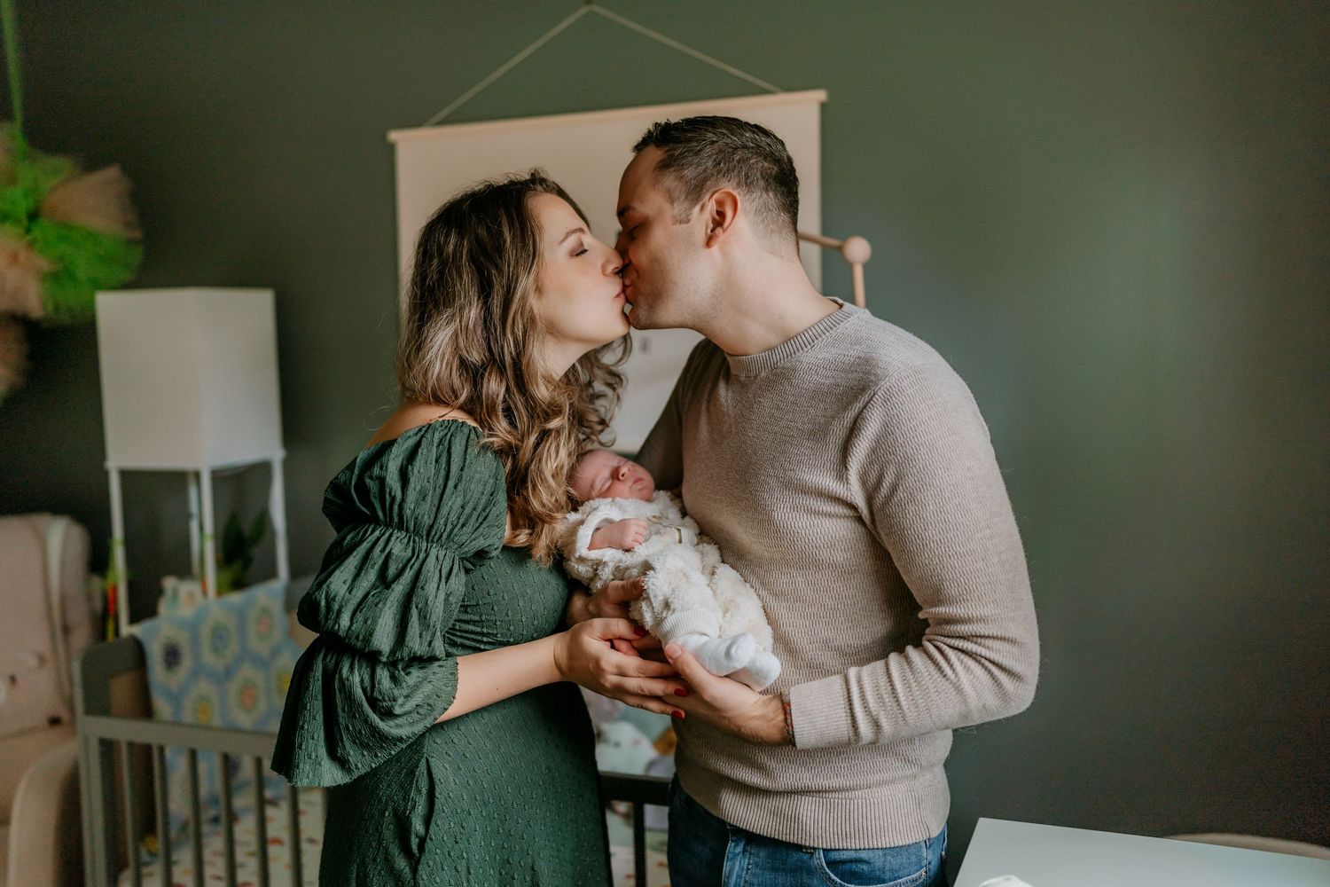 A couple shares an intimate moment while holding their small white dog in a cozy nursery setting.