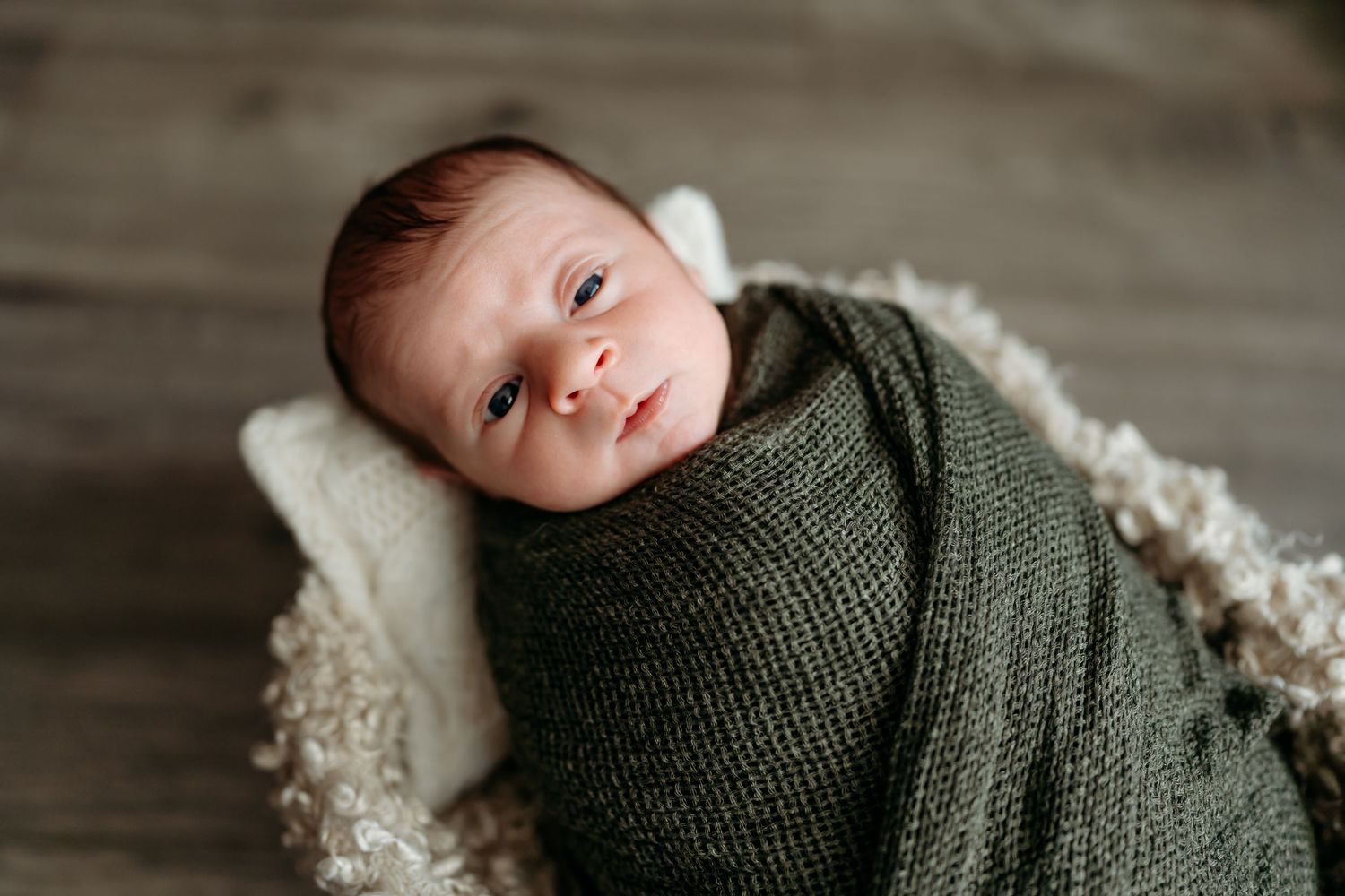 A newborn baby wrapped in a green knit blanket lying on a textured surface with wide eyes.