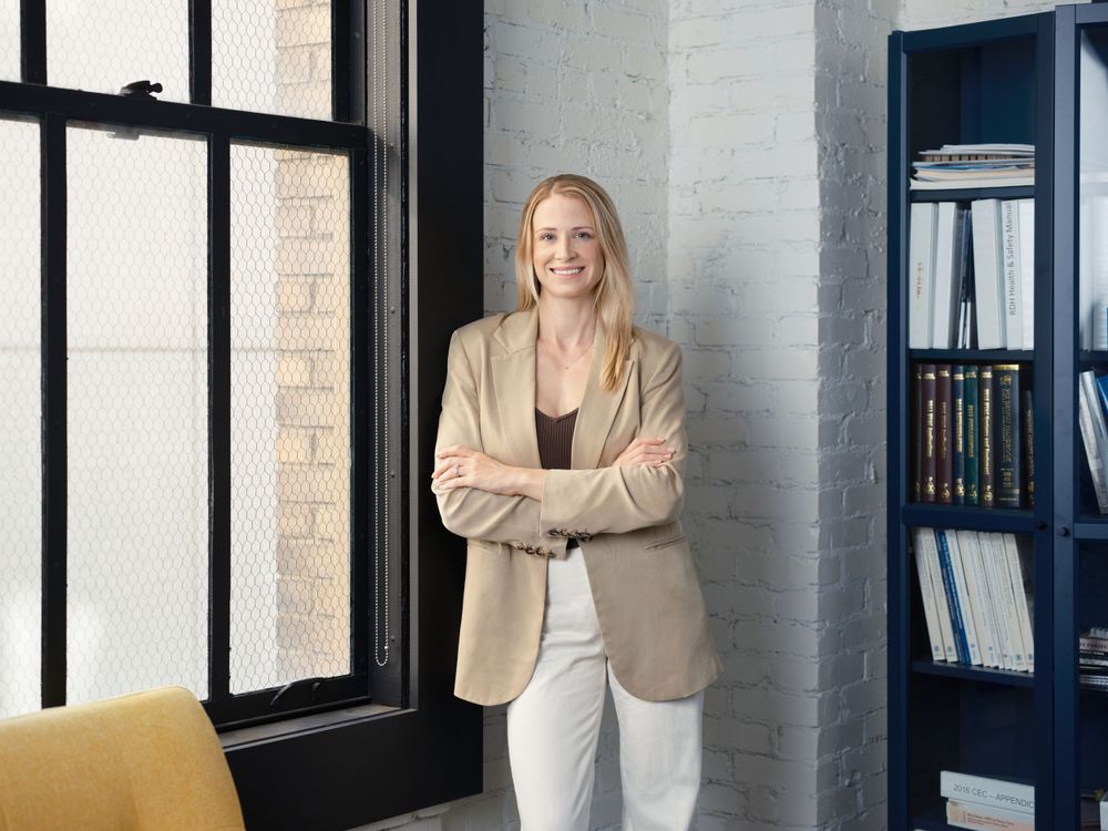 Professional headshot of a woman standing confidently in an industrial-style office with large windows — photographed in Oakland by Khoi Ly.
