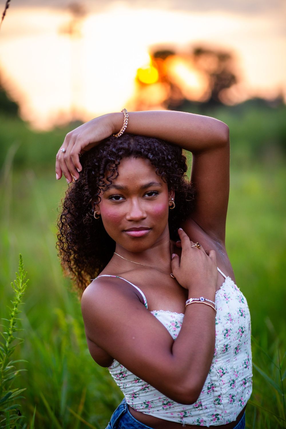 Beautiful portrait of a senior girl in a floral top against a sunset backdrop with tall grass and golden hour lighting in Marion, Iowa..