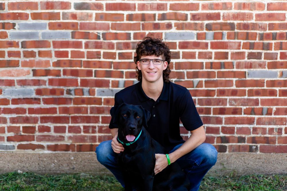 A senior boy crouches with a dog by a red brick wall background in Central City, Iowa.