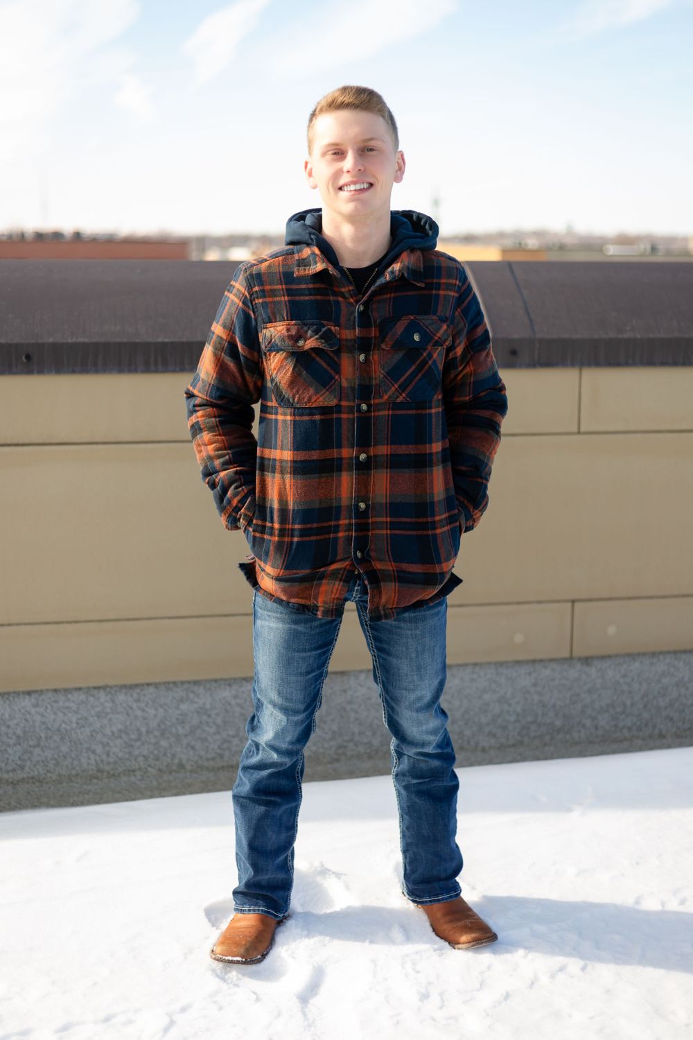 Senior boy stands in snow wearing a brown plaid flannel jacket, blue hoodie, jeans and work boots against a tan wall in Marion, Iowa.