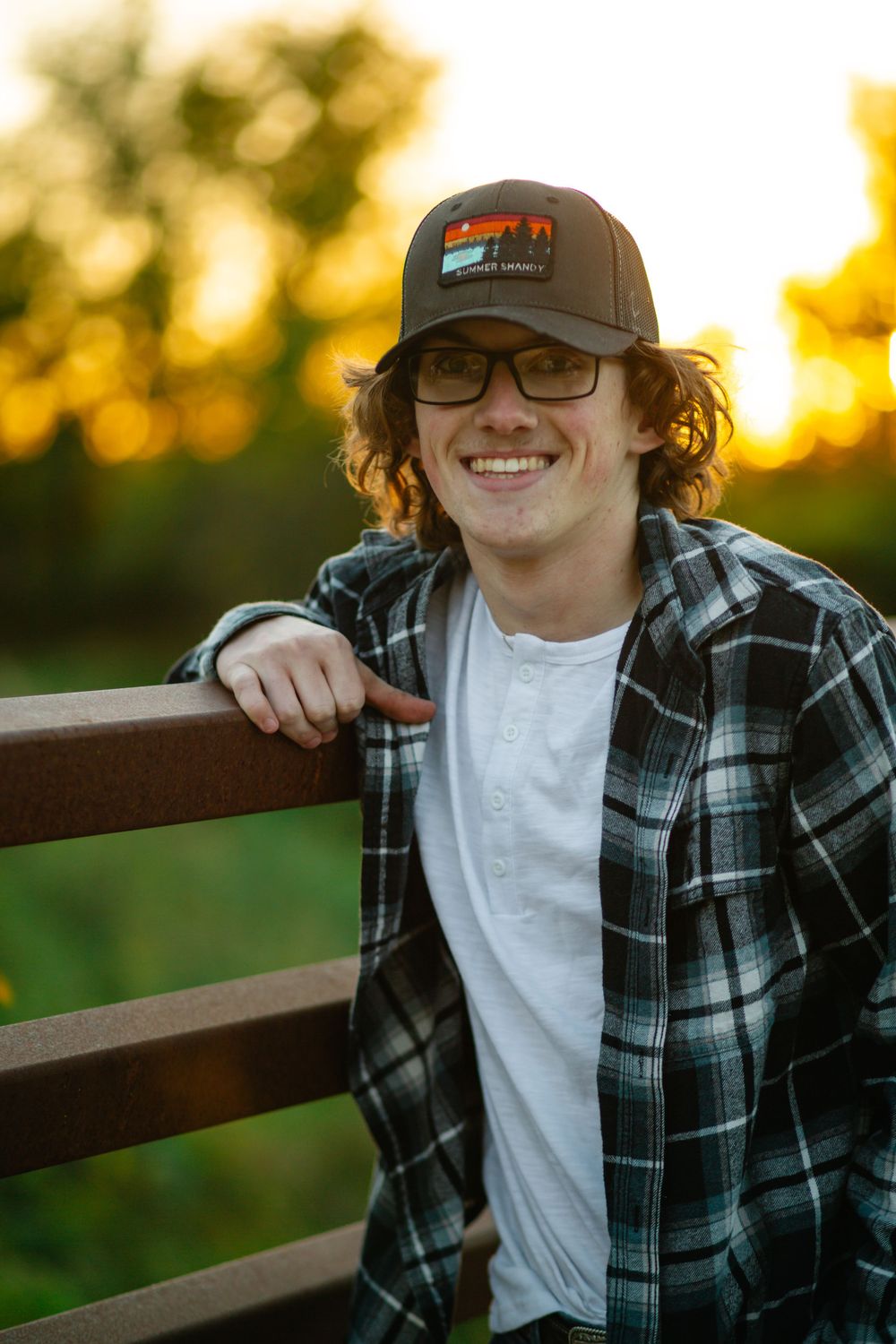 senior boy in a plaid flannel shirt and baseball cap leans against a wooden fence at sunset in Robins, Iowa.