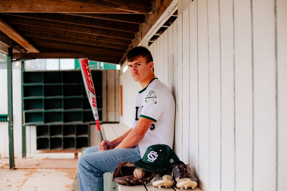 High school senior baseball player sits with bat in dugout while wearing casual jeans and white jersey uniform in Waterloo, Iowa.