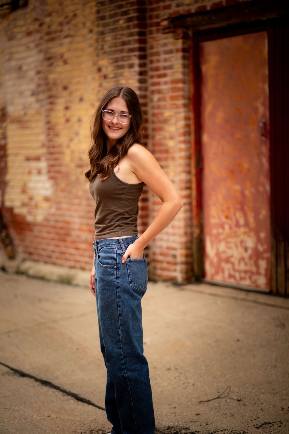 Portrait of a senior girl  in Anamosa, Iowa against brick wall with rustic door background wearing brown tank top and wide-leg jeans.