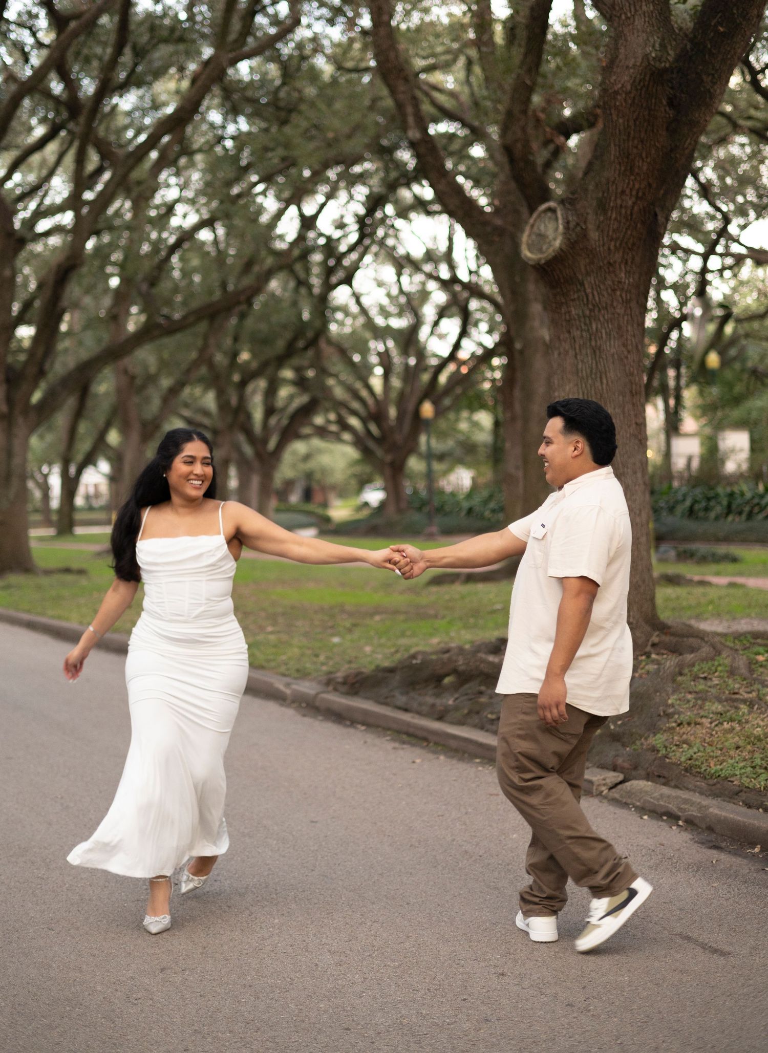 Two people in white outfits playfully dance together on a tree-lined path.