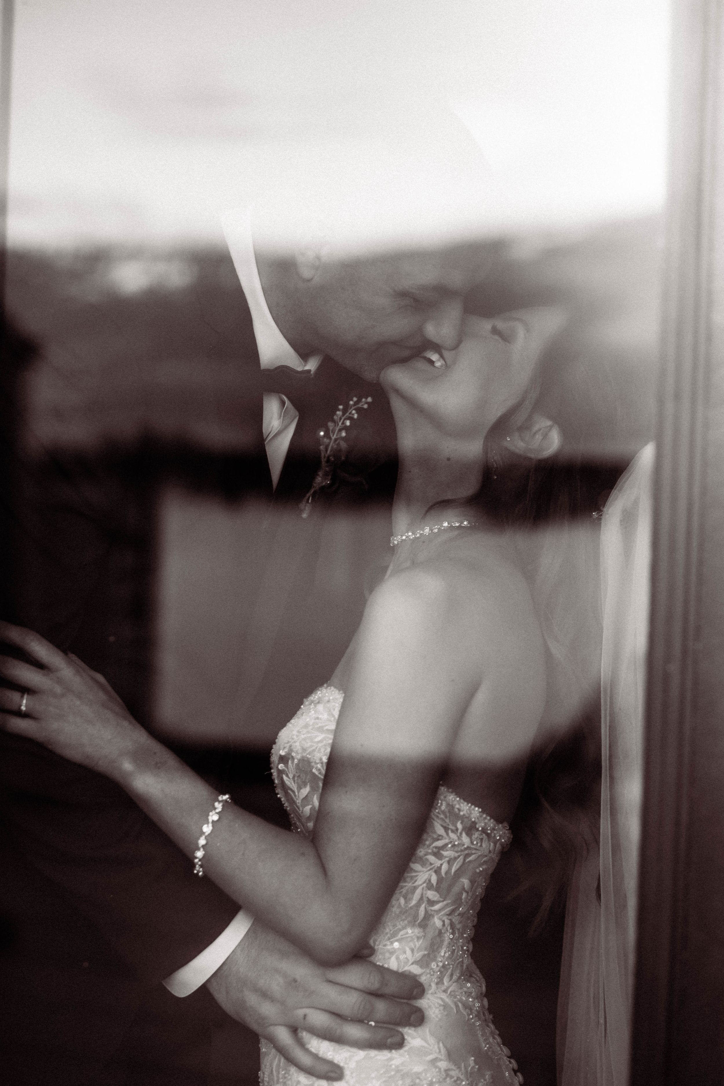 Artistic black and white wedding photo showing bride in lace dress with pearl bracelet looking through window reflection.