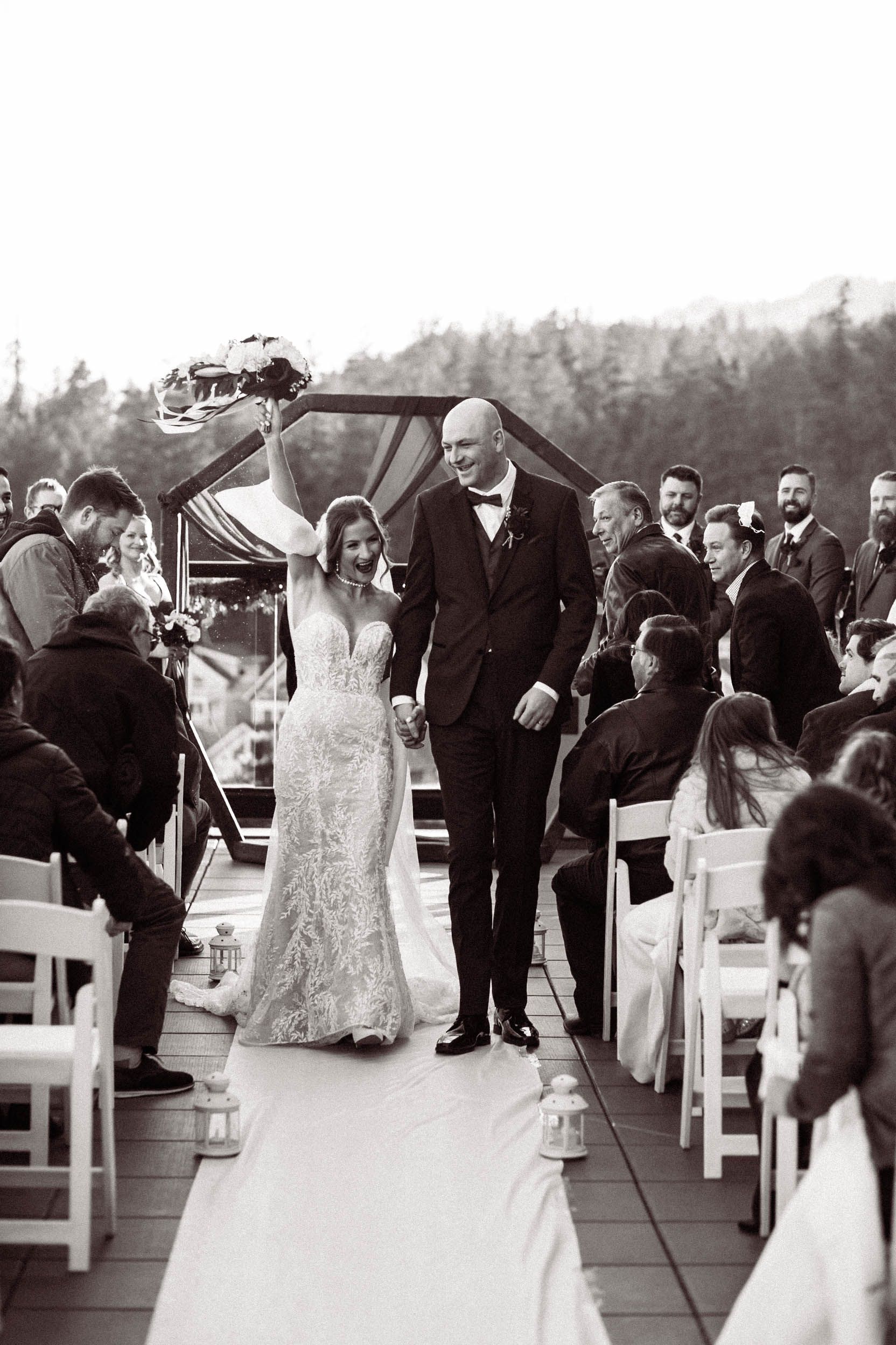 A black and white wedding photo captures the ceremony procession on a deck surrounded by guests seated in white chairs.