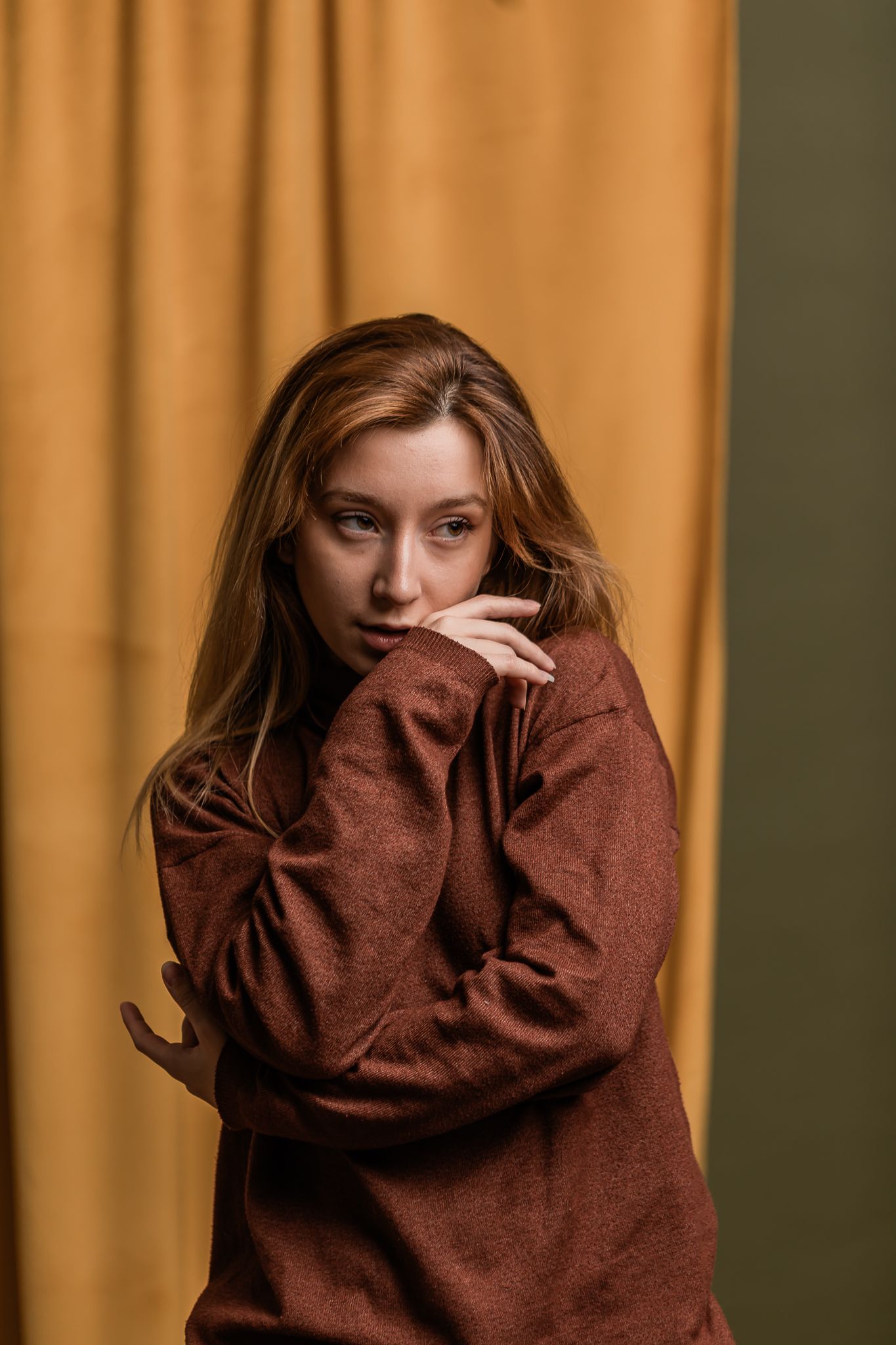 Aesthetic portrait against golden curtain backdrop features person in cozy brown sweater with flowing hair.