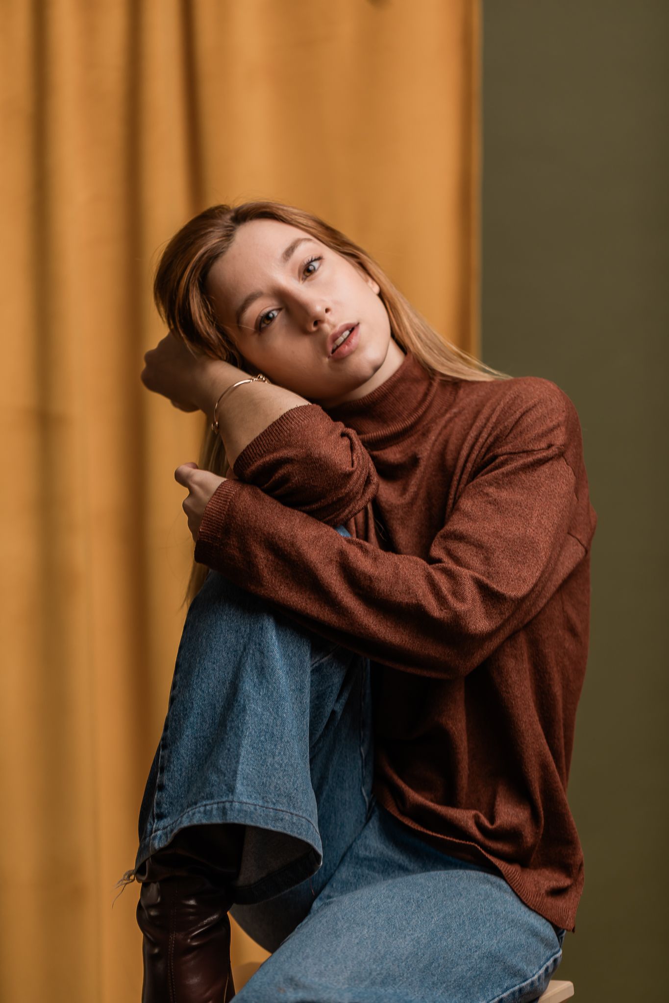 Model in brown turtleneck and jeans poses casually against orange curtain backdrop in artistic portrait.