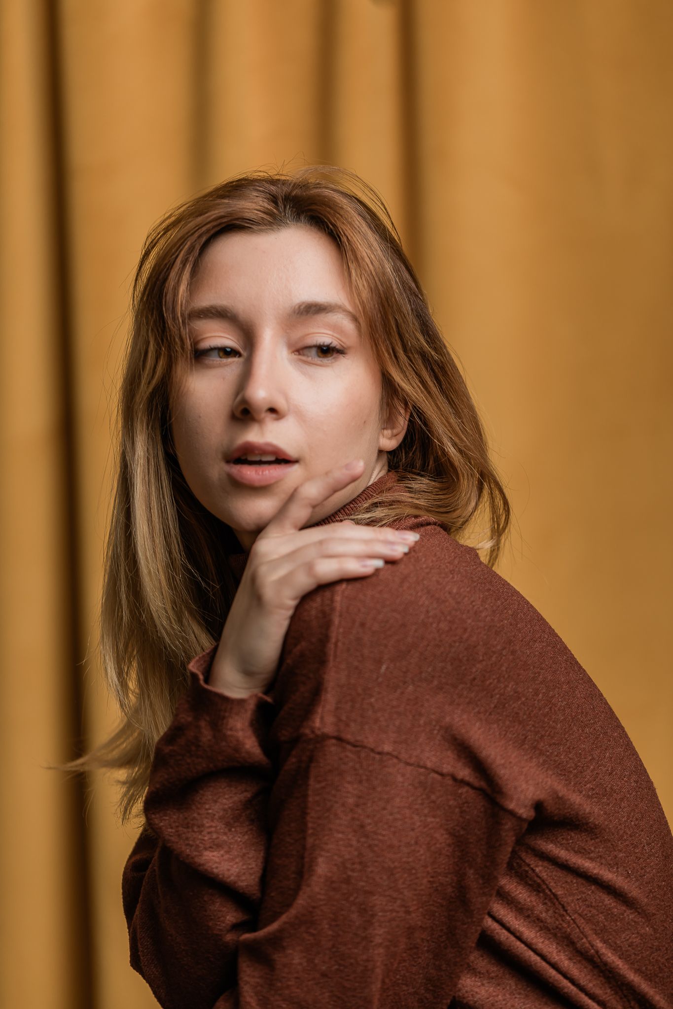 Portrait of a model in a brown sweater posing against an ochre curtain background with natural lighting and soft expression.
