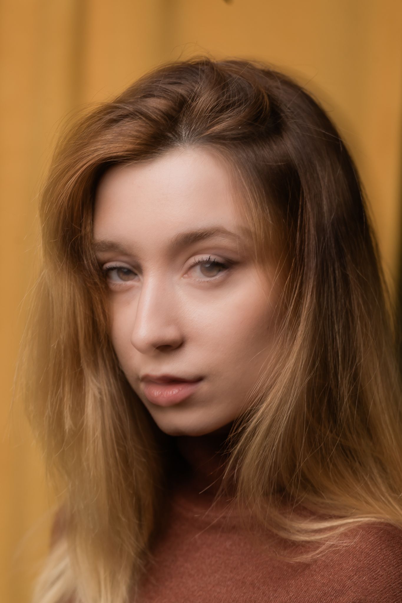 Portrait against orange background shows person with long brown hair wearing rusty brown sweater gazing to side.