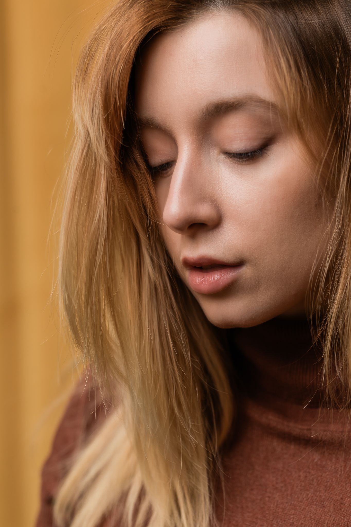 Close up portrait of woman with blonde hair and closed eyes wearing brown sweater against golden background.