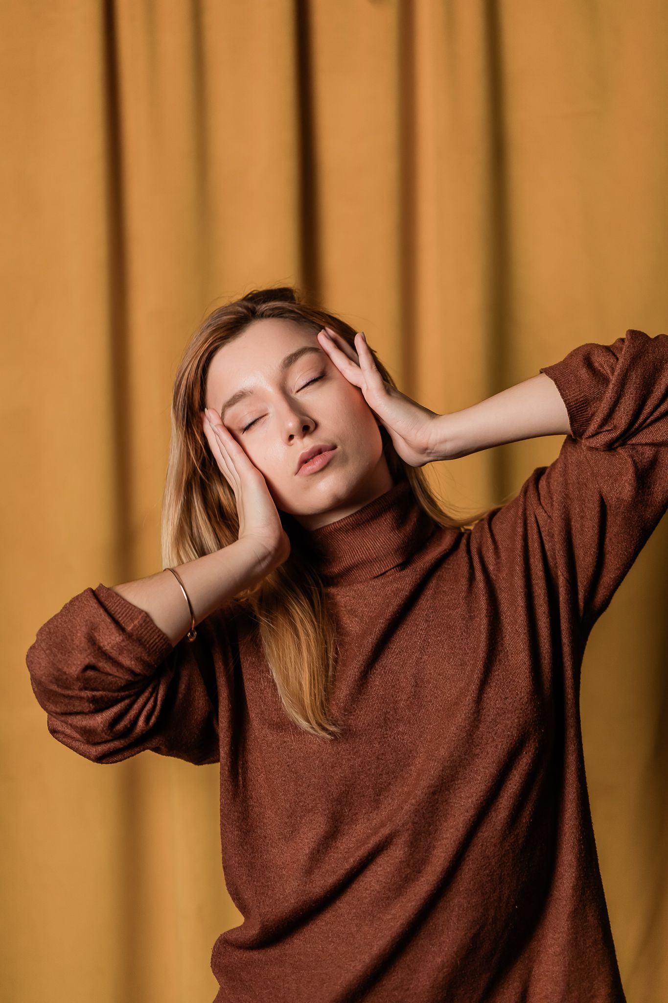 Person in a brown turtleneck sweater poses with hands on head against an orange backdrop with closed eyes.