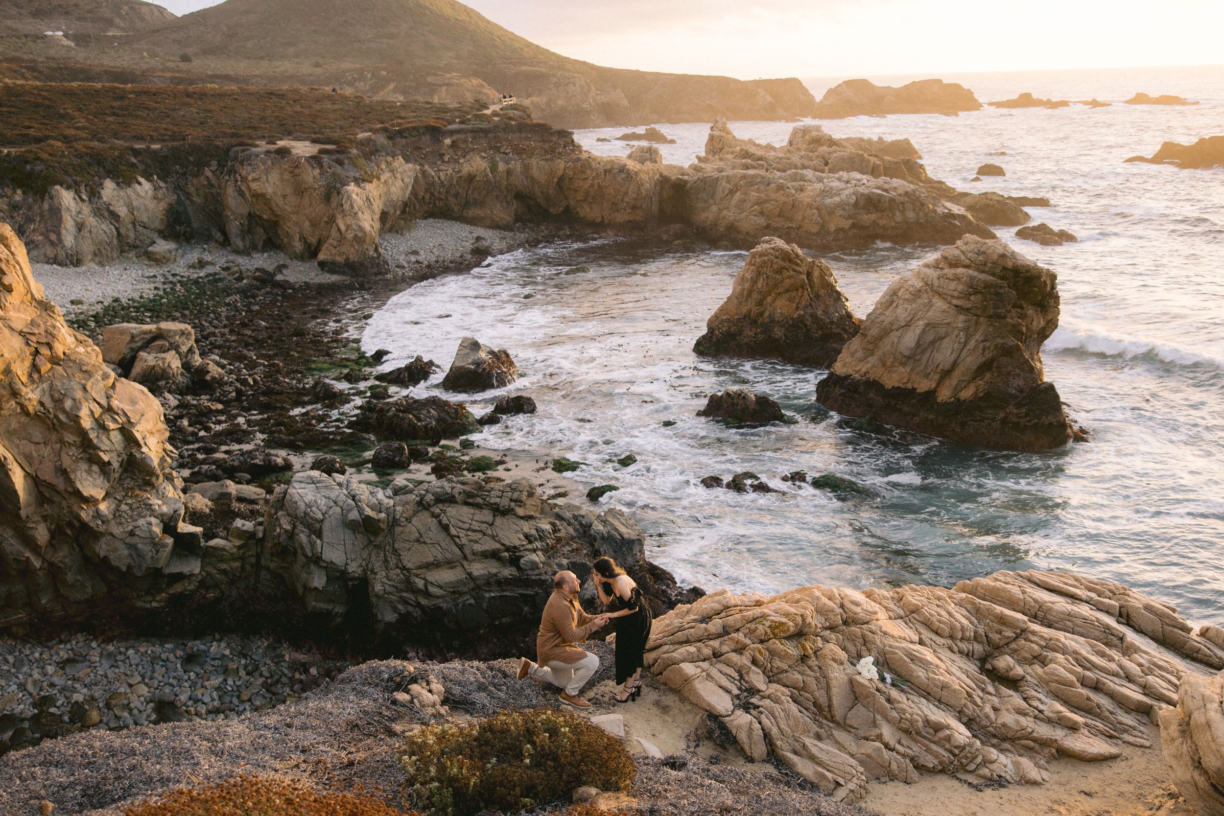Two people enjoying a sunset view from rocky coastal cliffs overlooking the dramatic Pacific Ocean shoreline.