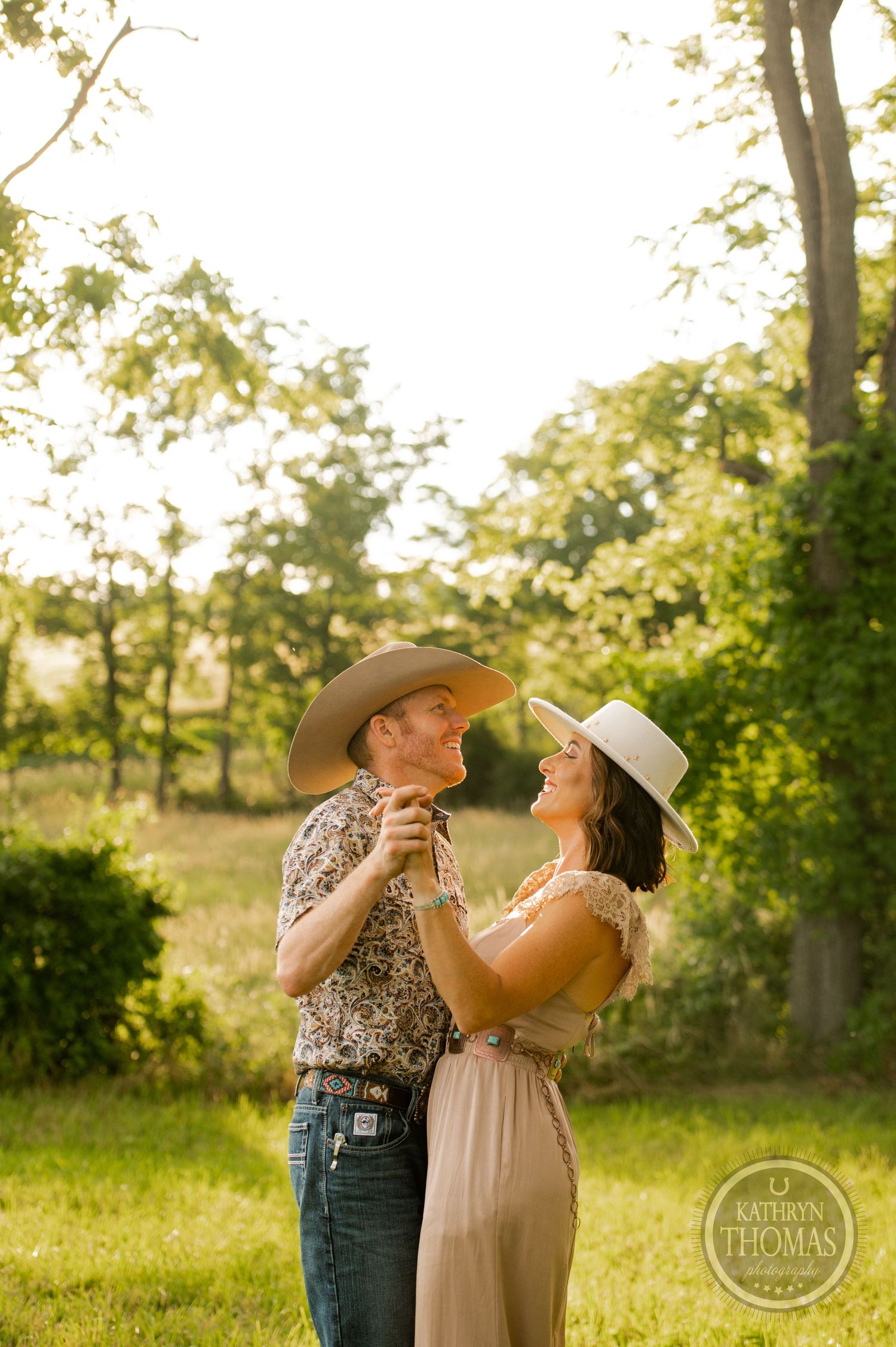 A couple in western attire sharing a playful moment in a sunlit meadow.